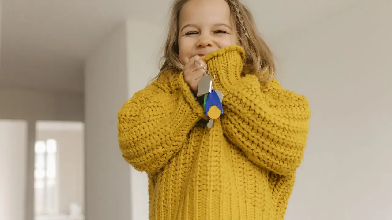 Ein fröhliches kleines Mädchen im gelben Pullover hält Schlüssel in der Hand. Die Frage "ab wann Kinder alleine lassen" beschäftigt Eltern oft, doch dieses Bild strahlt Unbeschwertheit aus.