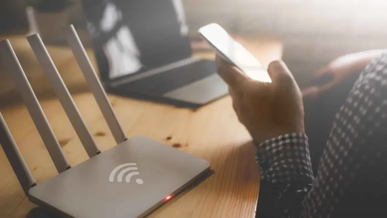 Router Wi-Fi, smartfon w dłoni, laptop na drewnianym biurku.