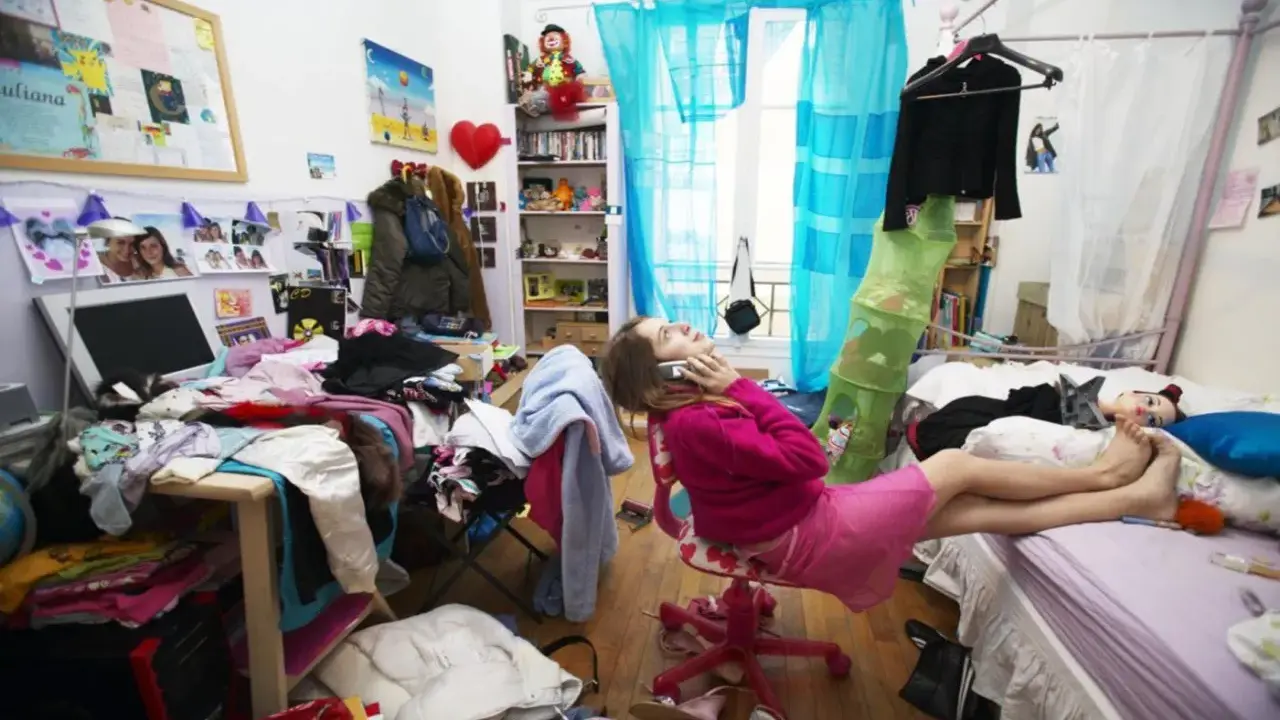 Chica en su habitación desordenada, hablando por teléfono, rodeada de ropa y objetos esparcidos.