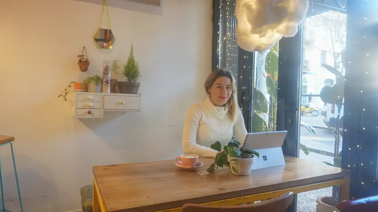 Kobieta pracuje na laptopie w kawiarni, ciesząc się kawą i roślinami. To idealne miejsce na dodatkową pracę zdalną.