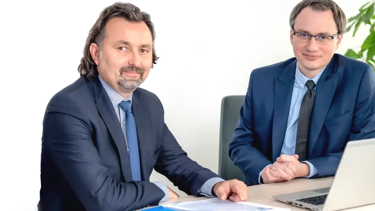Dwóch mężczyzn w garniturach siedzi przy biurku z dokumentami i laptopem.