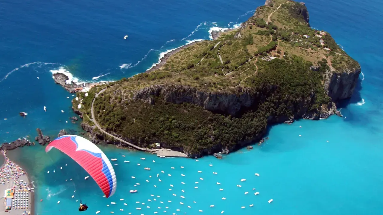 Parapendio su un mare turchese vicino a isole calabresi, con una spiaggia affollata e tante barche.