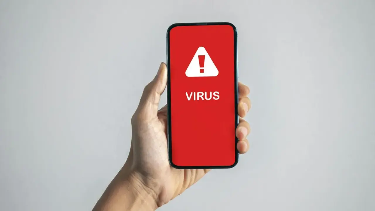 En hand håller en mobil med en röd skärm som visar en varningstriangel och texten "VIRUS". En påminnelse om hur vet man om man har virus.