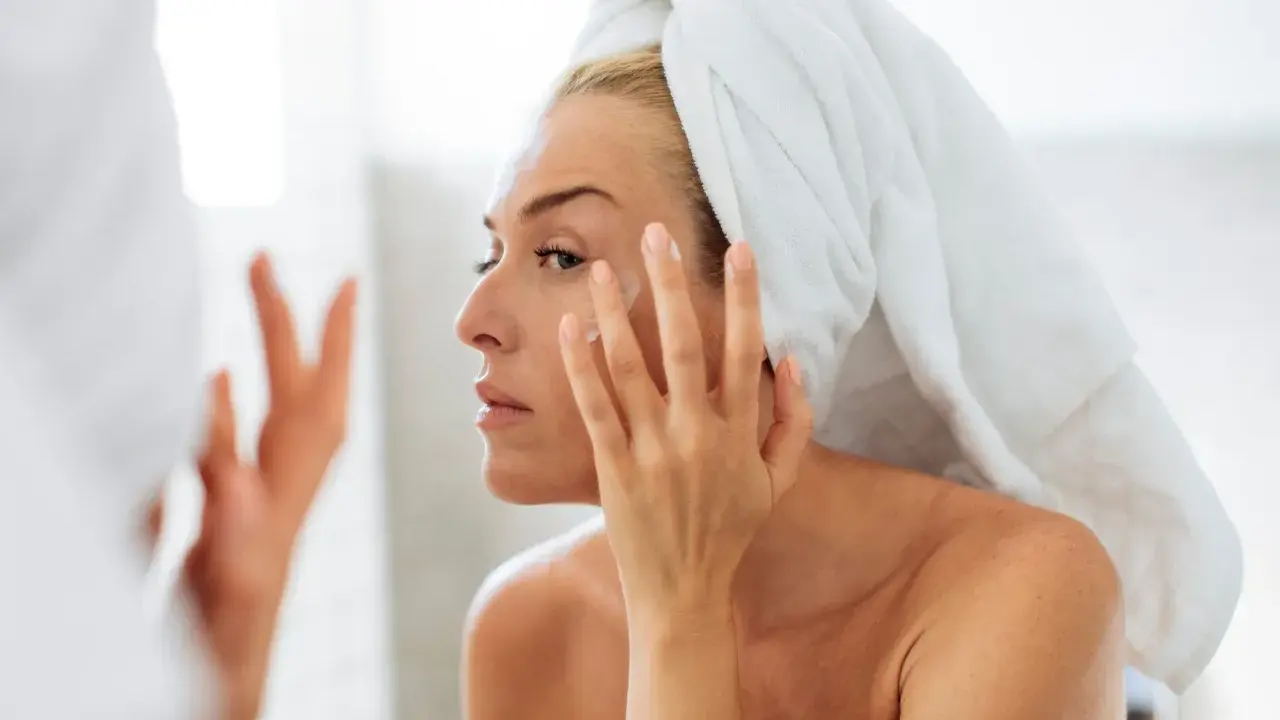 Une femme, serviette sur la tête, applique une crème sur son visage. Un geste essentiel pour prendre soin de sa peau en hiver.