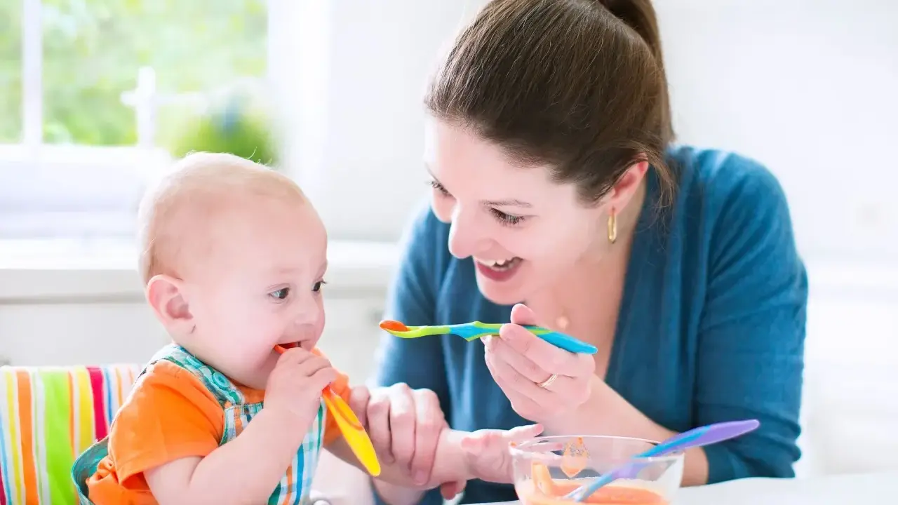 Uśmiechnięta mama karmi łyżeczką 7 miesięczne dziecko, które samo próbuje jeść.