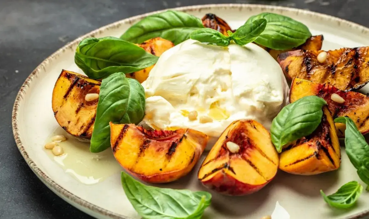 Burrata z grillowanymi brzoskwiniami, bazylią i orzeszkami piniowymi na talerzu.