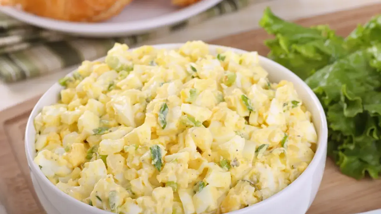 Pyszna sałatka z jajkiem, idealna na śniadanie lub lunch. Podana w białej misce, obok świeżych liści sałaty i chrupiących croissantów.