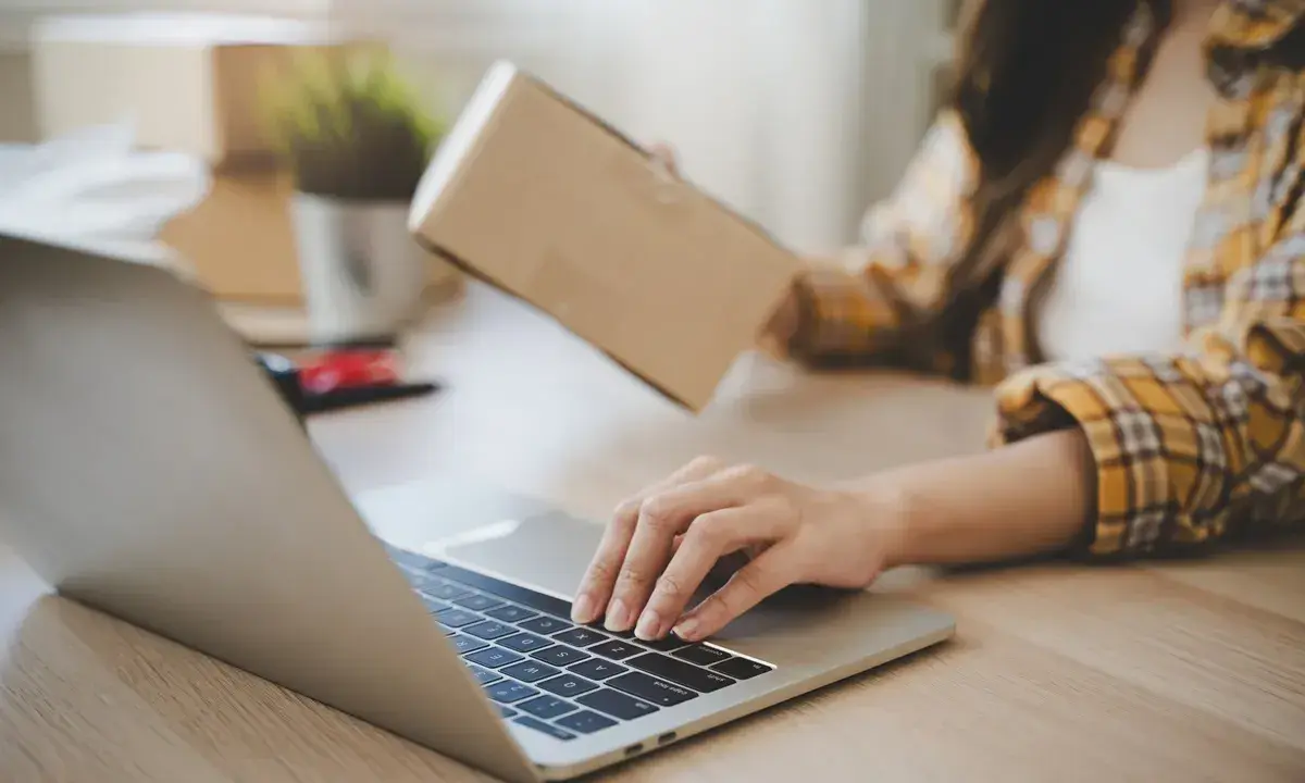Osoba pakuje paczkę, sprawdzając na laptopie, kto ponosi koszty zwrotu towaru.