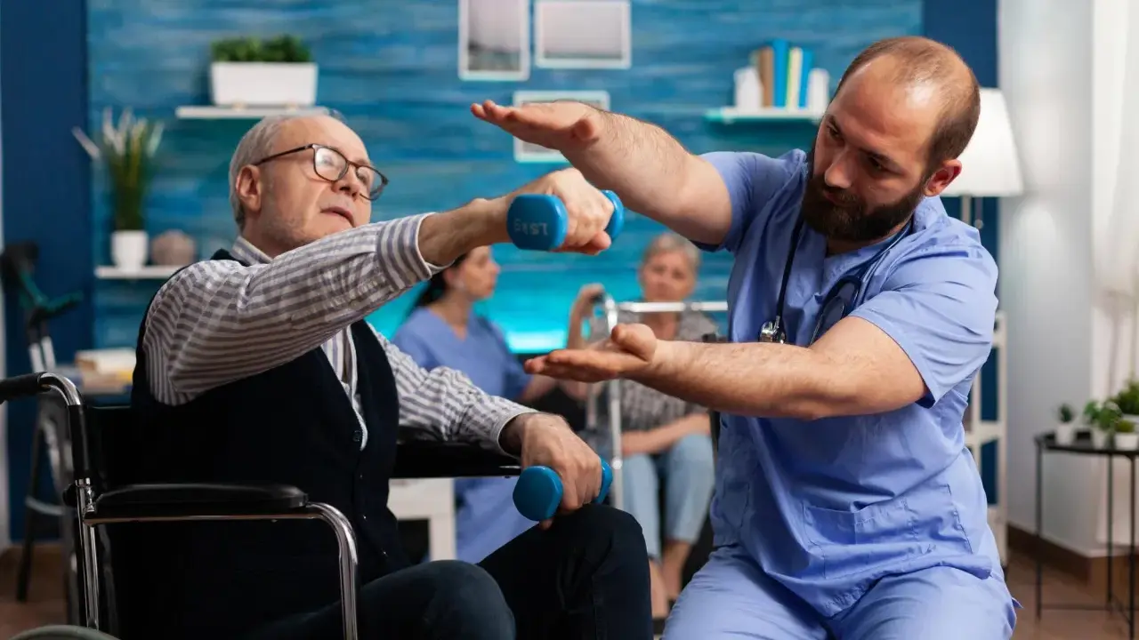 Starszy mężczyzna w wózku wykonuje ćwiczenia z hantlem pod okiem fizjoterapeuty. To kluczowy element rehabilitacji ręki po udarze.