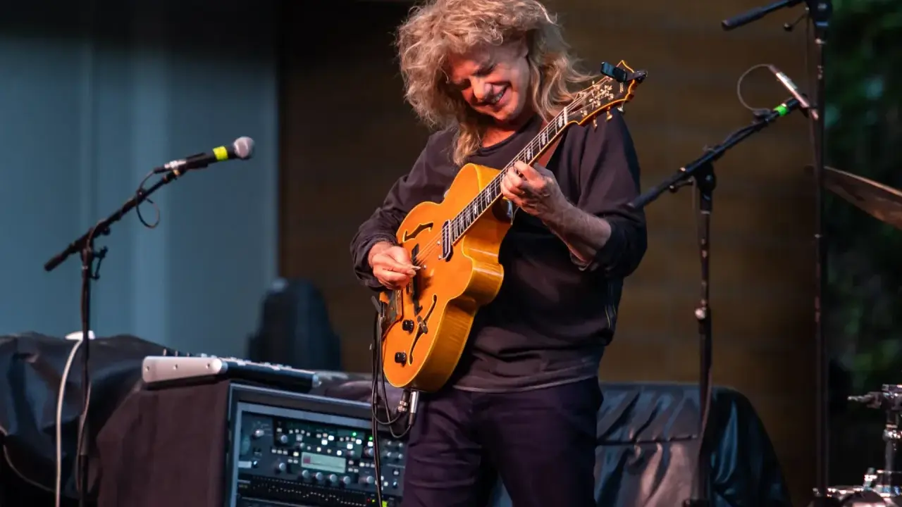 Muzyk z burzą blond włosów gra na gitarze, tworząc wspaniały śpiew w duecie z niewidocznym wokalistą.