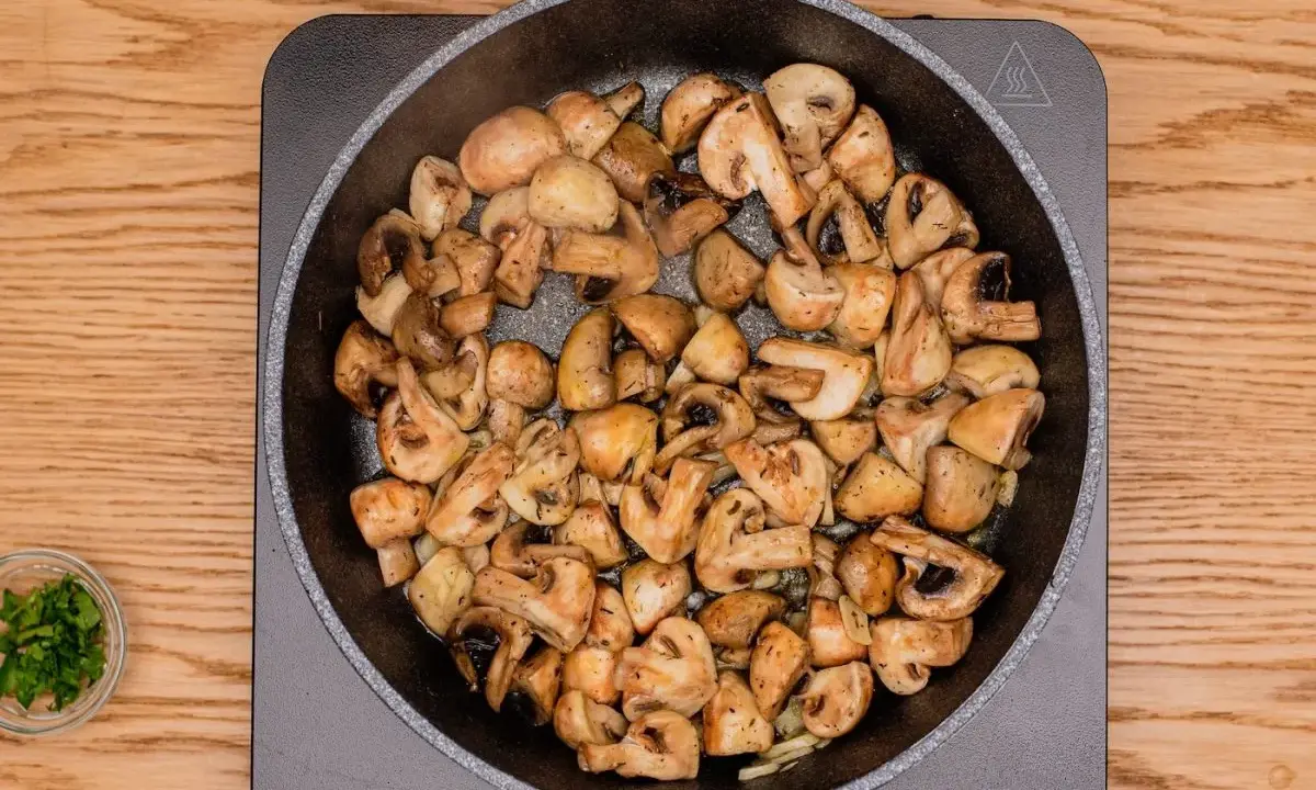 Champiñones al ajillo dorándose en una sartén, con perejil picado en un cuenco al lado.