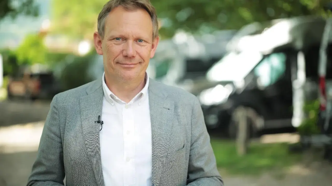 Johannes Sturm, ein Mann mittleren Alters mit freundlichem Lächeln, trägt ein Sakko und ein Hemd. Im Hintergrund sind Bäume und Fahrzeuge zu sehen.