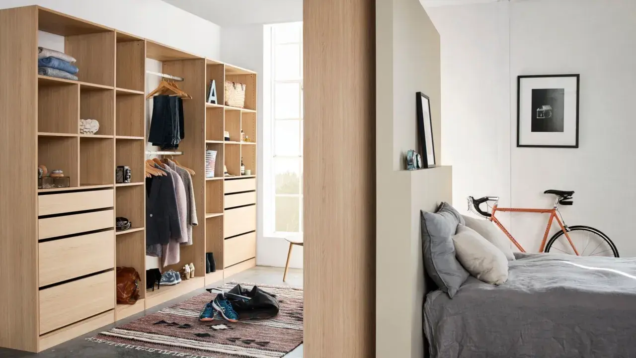 Un armario moderno y funcional, ideal para aprender como organizar un armario. Ropa colgada, cajones y estantes ordenados, con una bicicleta naranja y una cama al fondo.