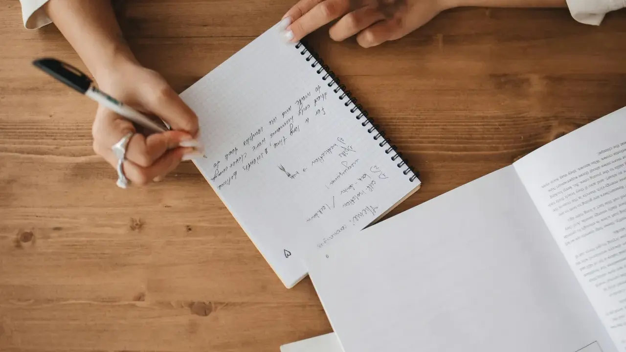 A person's hands are writing notes in a notebook. The notes mention "Theme: coronavirus", "self-isolation", and "lockdown".