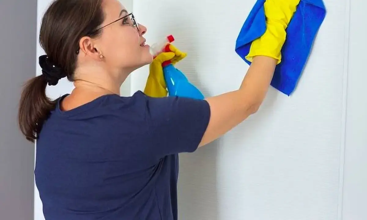Mujer con guantes amarillos y paño azul limpia puerta lacada en blanco con spray.