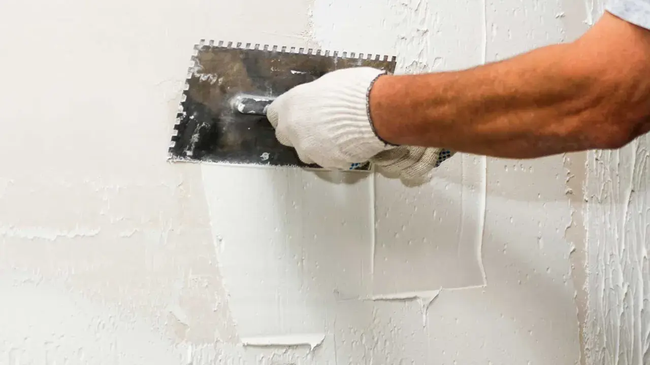 Mano enguantada aplicando estuco a una pared con una llana dentada. El proceso de estucar una pared requiere precisión.