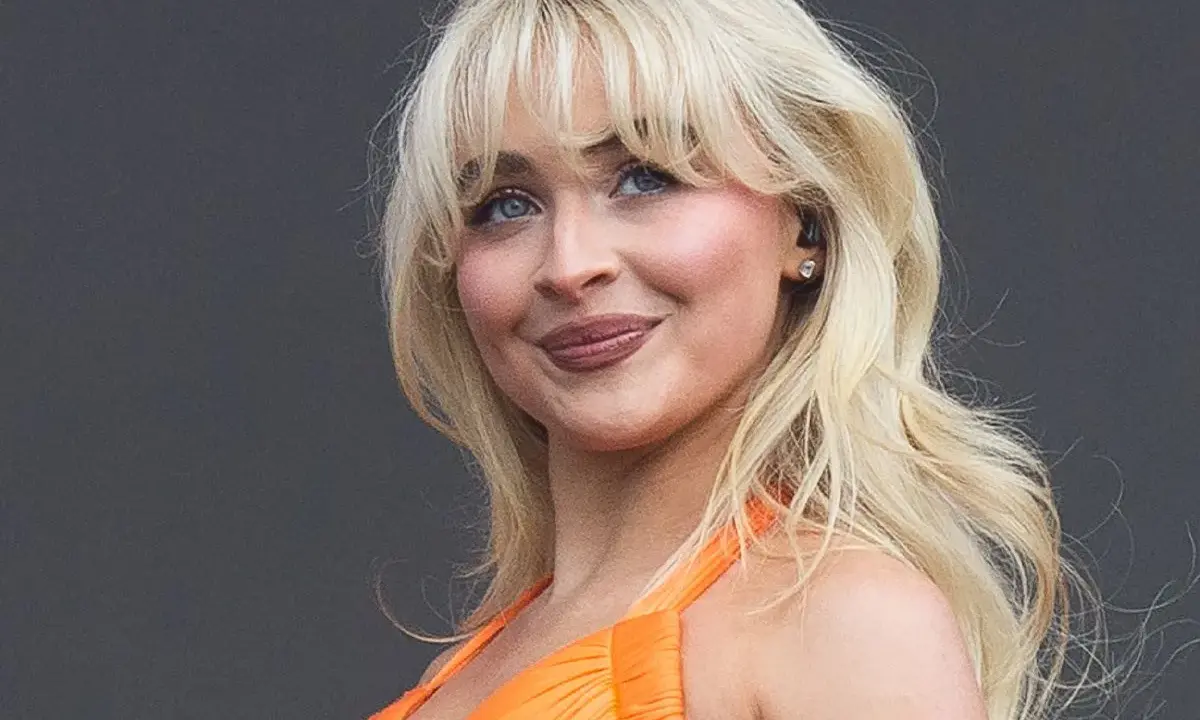 Blonde Frau mit voluminösen Blow-out-Haaren und einem strahlenden Lächeln in einem orangefarbenen Kleid.