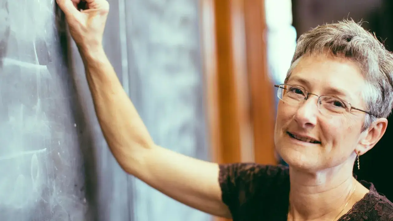 Lehrerin schreibt an Tafel. Was verdient ein Gymnasiallehrer? Sie lächelt wissend.