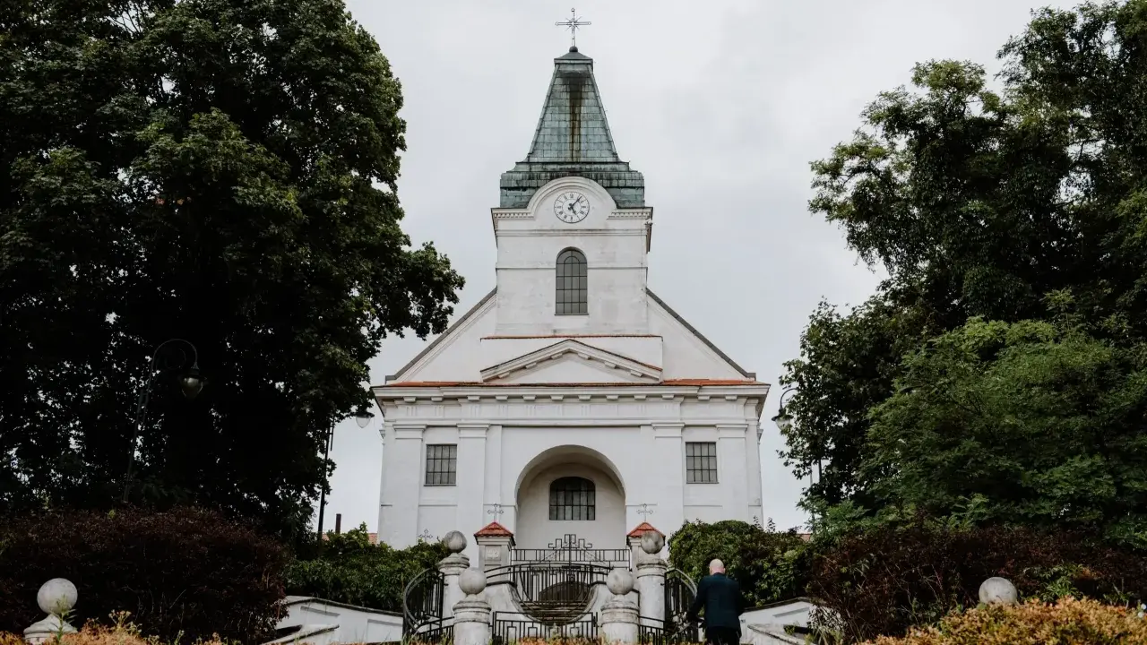 Biały kościół z zegarem na wieży, otoczony drzewami. Zastanawiasz się, jak sprawdzić do której parafii należę?