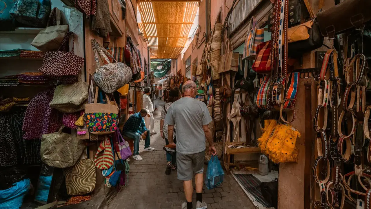 Bunte Taschen und Gürtel hängen in einem engen Gang des Marrakesch Basars. Ein Mann geht mit einer blauen Tüte durch das bunte Treiben.
