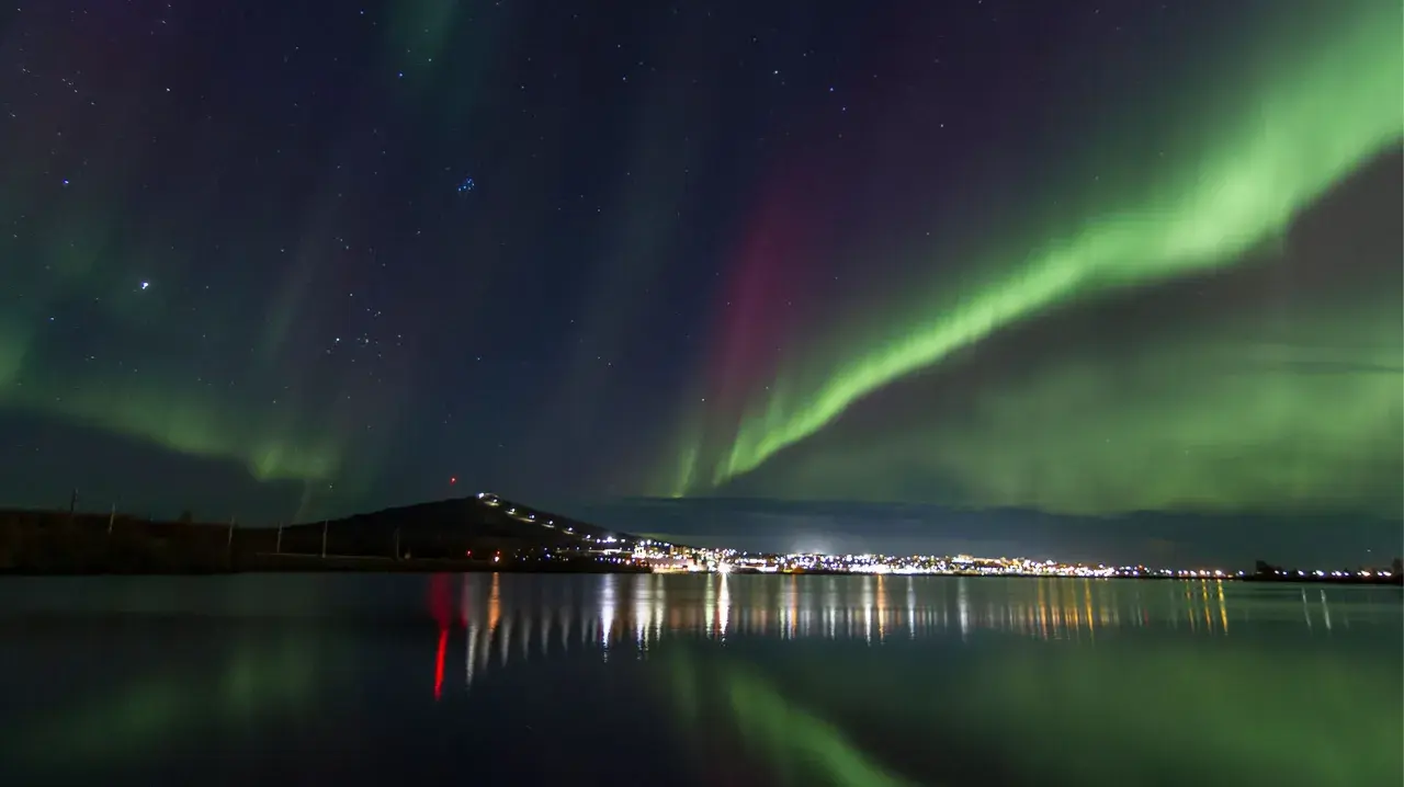 Gröna och lila norrsken dansar över en stjärnklar himmel ovanför Kiruna. Stadens ljus speglas i det lugna vattnet.