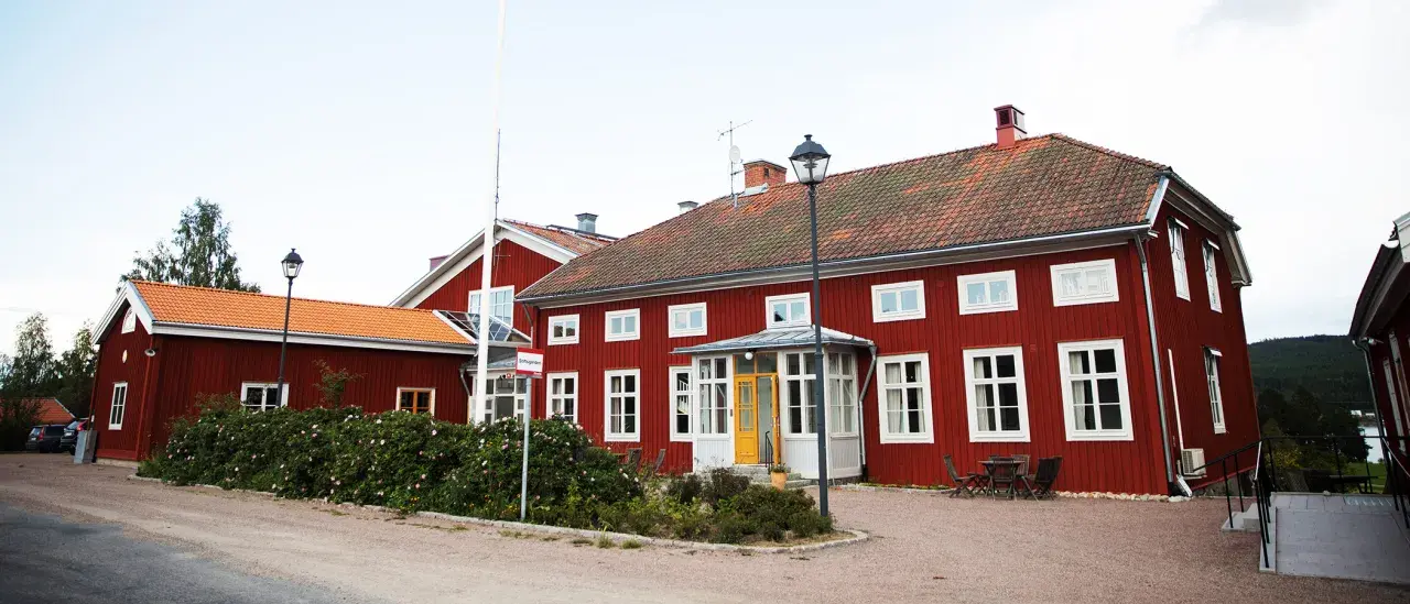 Röda trähus med vita fönster och tegeltak. En skylt visar "Stiftsgården Undersvik". Vackert beläget med natur och vatten i bakgrunden.