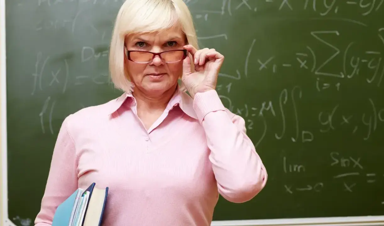 Nauczycielka z okularami i książkami przy tablicy z matematycznymi wzorami. Czas na zasłużone emerytury dla nauczycieli.