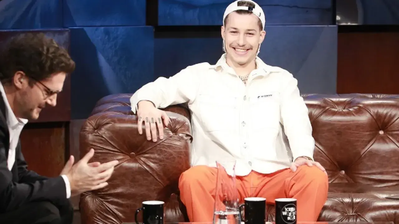 Zabson, z uśmiechem na twarzy, siedzi na kanapie w studiu. Obok niego siedzi mężczyzna w okularach.