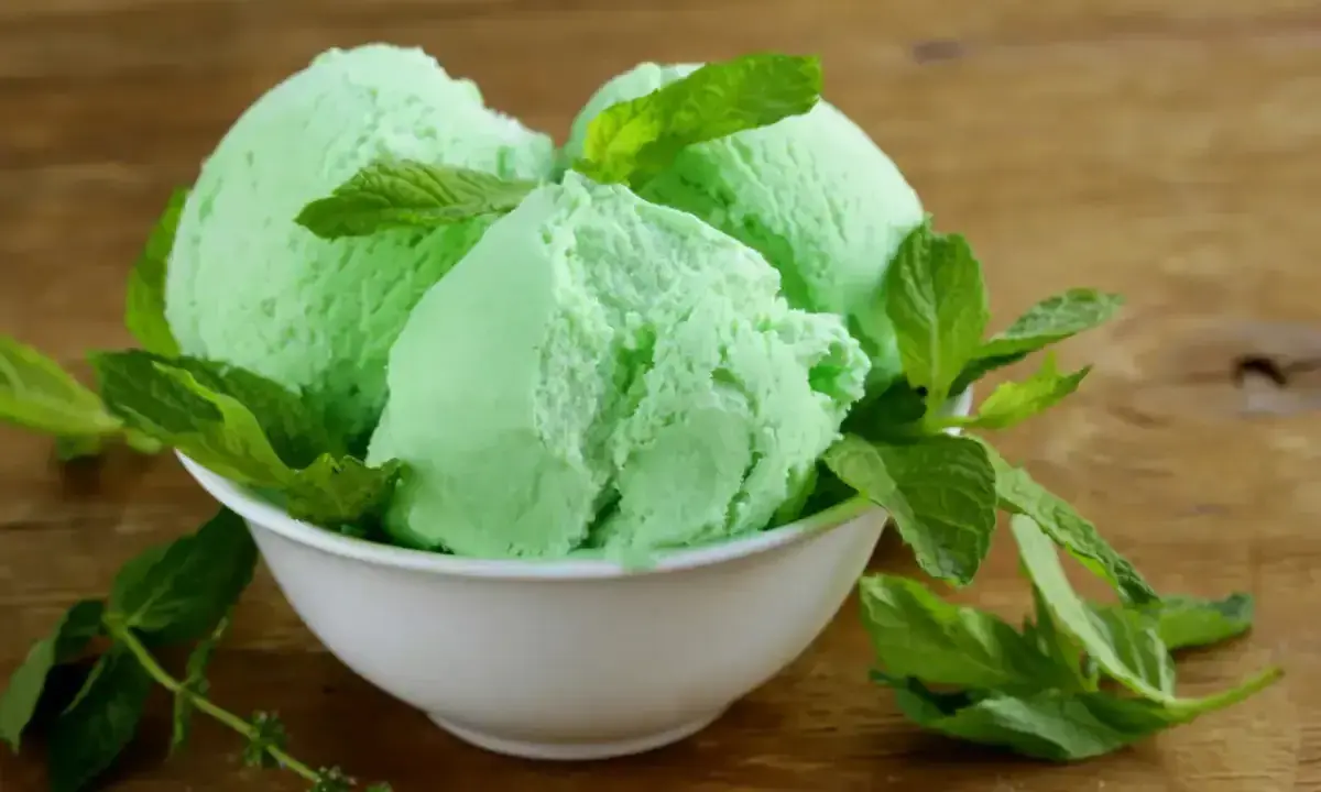Trois boules de glace verte menthe, rappelant la saveur de la glace Get 27, garnies de feuilles de menthe fraîche dans un bol blanc sur fond de bois.