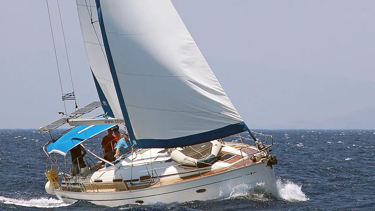 Voile blanche gonflée par le vent, le voilier avance sur la mer bleue. La bosse de ris sur la grand-voile est visible, signe d'une navigation par vent fort.