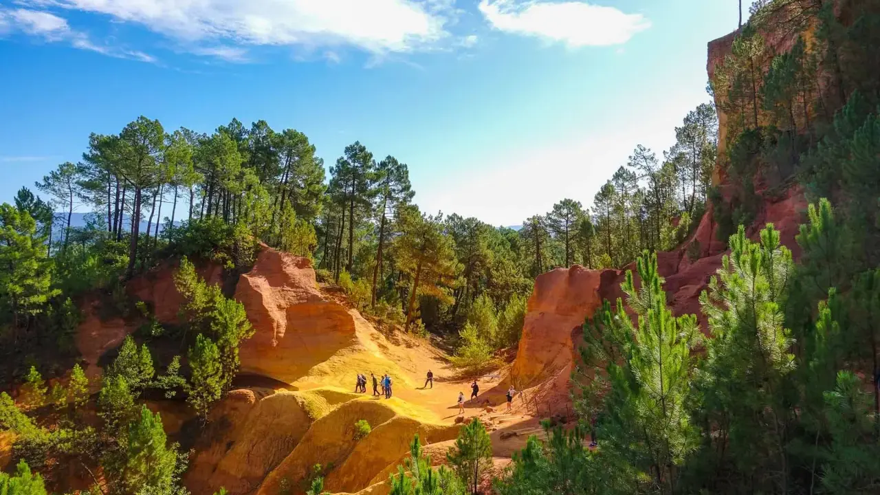 Des randonneurs explorent le sentier des ocres, un paysage spectaculaire aux falaises rouges et jaunes, bordé de pins verts sous un ciel bleu.