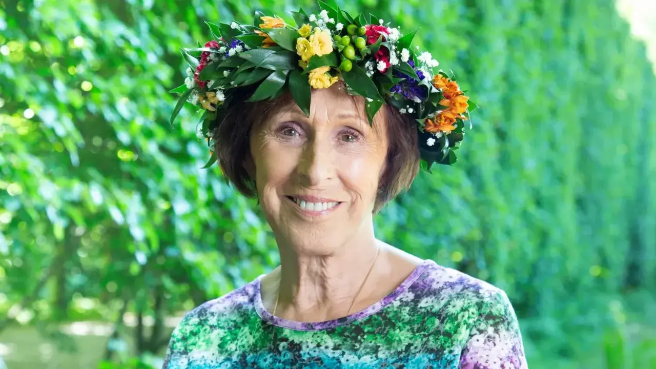Eva Rydberg ler glatt med en blomsterkrans på huvudet, omgiven av grönska.