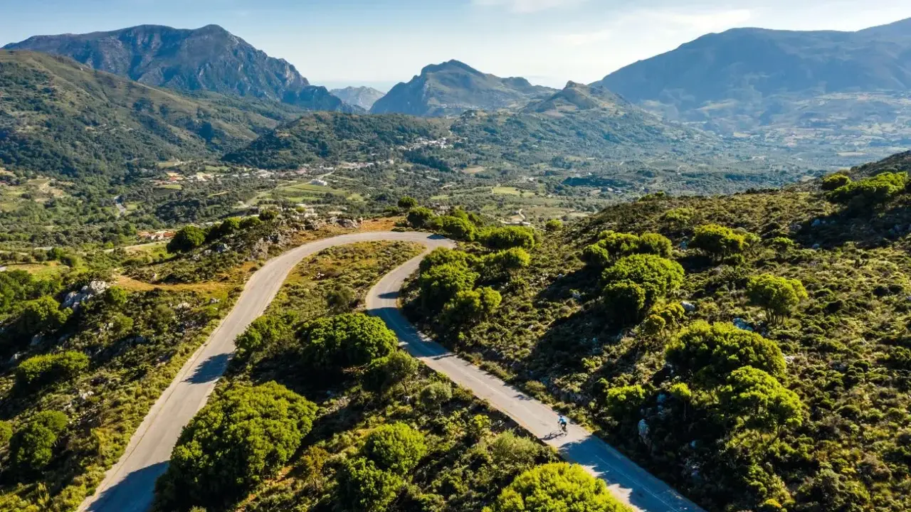 Ein Rennradfahrer auf Kreta genießt die kurvenreiche Straße durch eine grüne Berglandschaft.