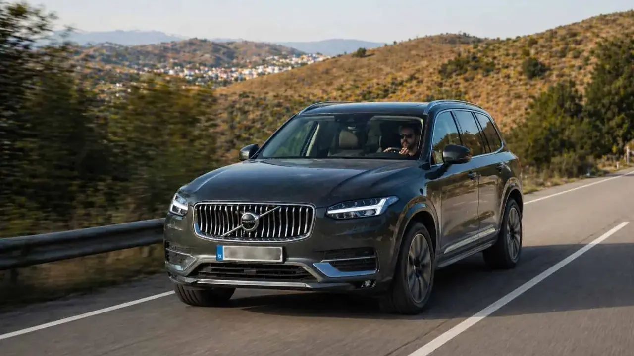 Volvo XC90 oscuro, uno de los suv mas fiables, circulando por una carretera rural con colinas verdes de fondo.