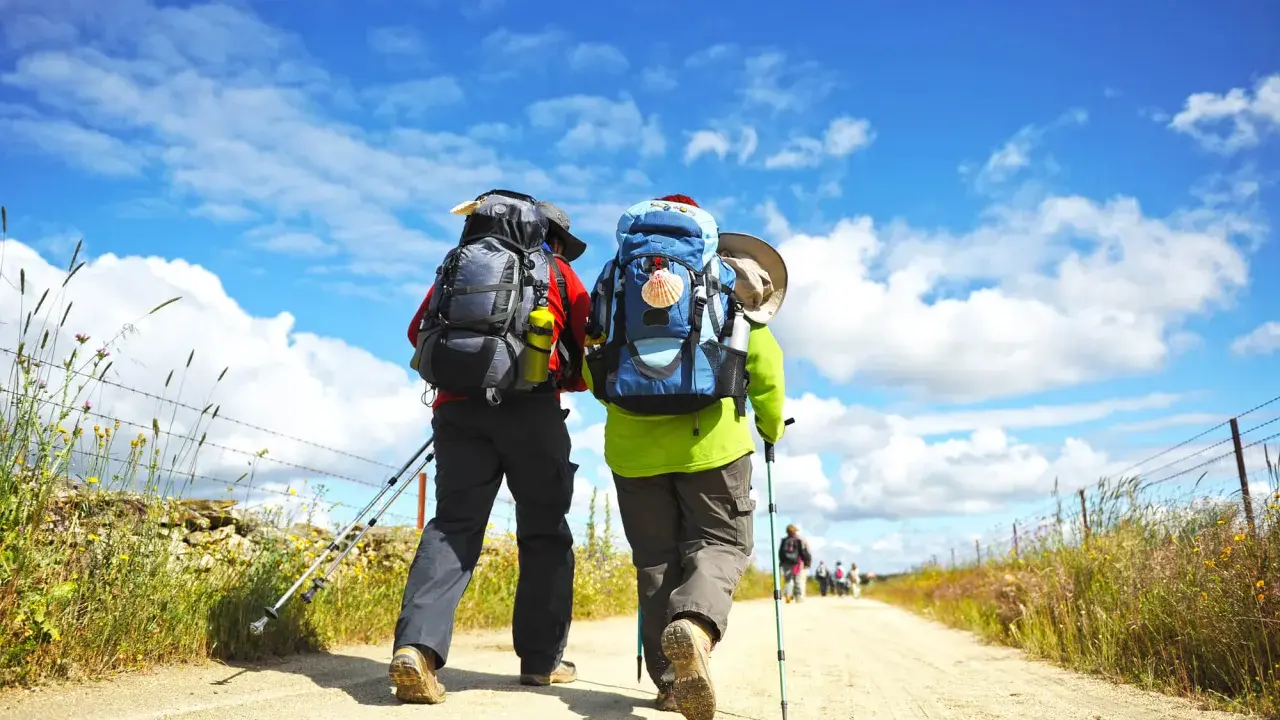 Två vandrare med ryggsäckar går längs en grusväg under en blå himmel med moln. En perfekt dag för pilgrimsvandring i Sverige.