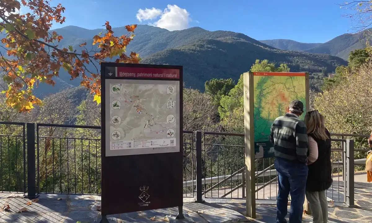 Pareja observa mapa de senderismo en los valles oriental de Montseny. El paisaje montañoso y el follaje otoñal crean un entorno pintoresco.