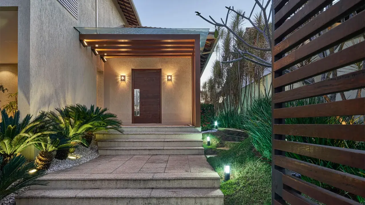 Un portico moderno con porta in legno scuro, illuminazione soffusa e piante rigogliose. Un esempio di come abbellire un portico con stile e natura.
