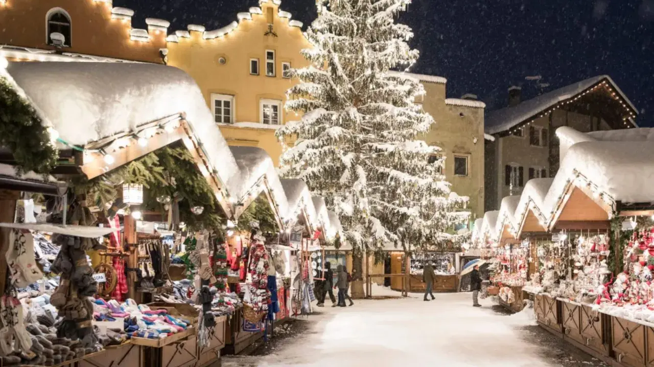 Mercatini di Natale nel Nord Italia, con bancarelle innevate, un grande albero illuminato e case decorate. Magia invernale.