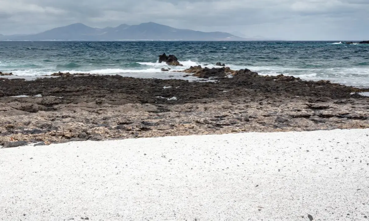 Arena blanca y rocas volcánicas en la playa Palomitas, Fuerteventura. El mar azul choca contra las rocas, con montañas al fondo.