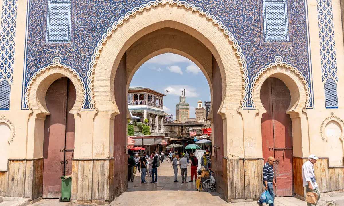 Prachtvolles Tor in Fes Stadt mit blau-goldenen Mustern. Menschen beleben den Platz dahinter.