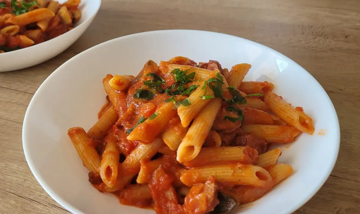 Penne z sosem pomidorowym i kawałkami kiełbasy, posypane natką pietruszki. Pyszny makaron z kiełbasą na białym talerzu.