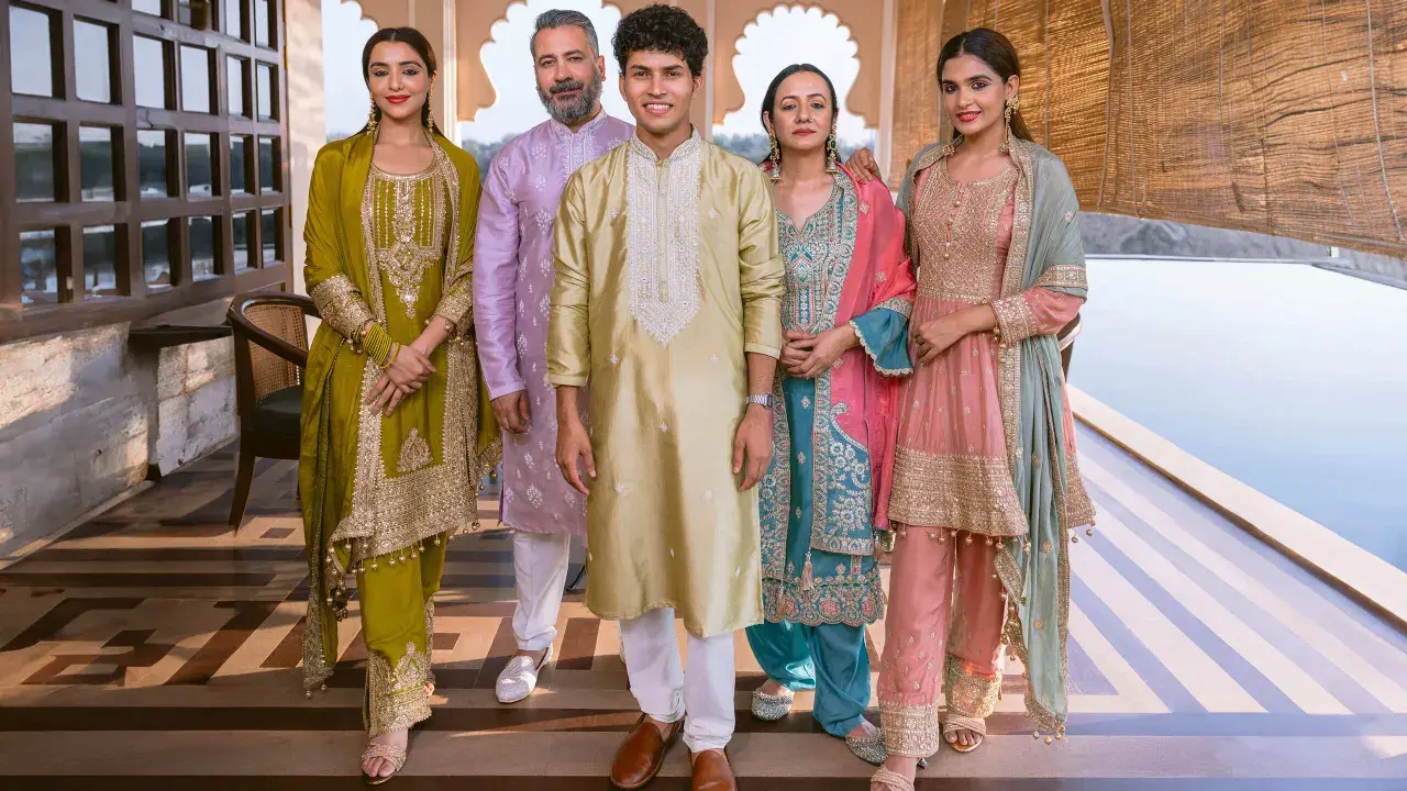 A family poses in vibrant traditional clothing from India. The women wear ornate salwar kameez sets in olive green, teal, and pink, while the men are dressed in elegant kurtas.