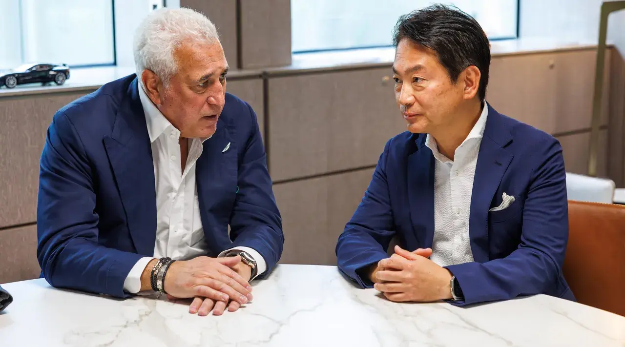 Koji Watanabe conversa con un hombre mayor, ambos con trajes azules. Un modelo de coche deportivo negro se ve en el fondo.