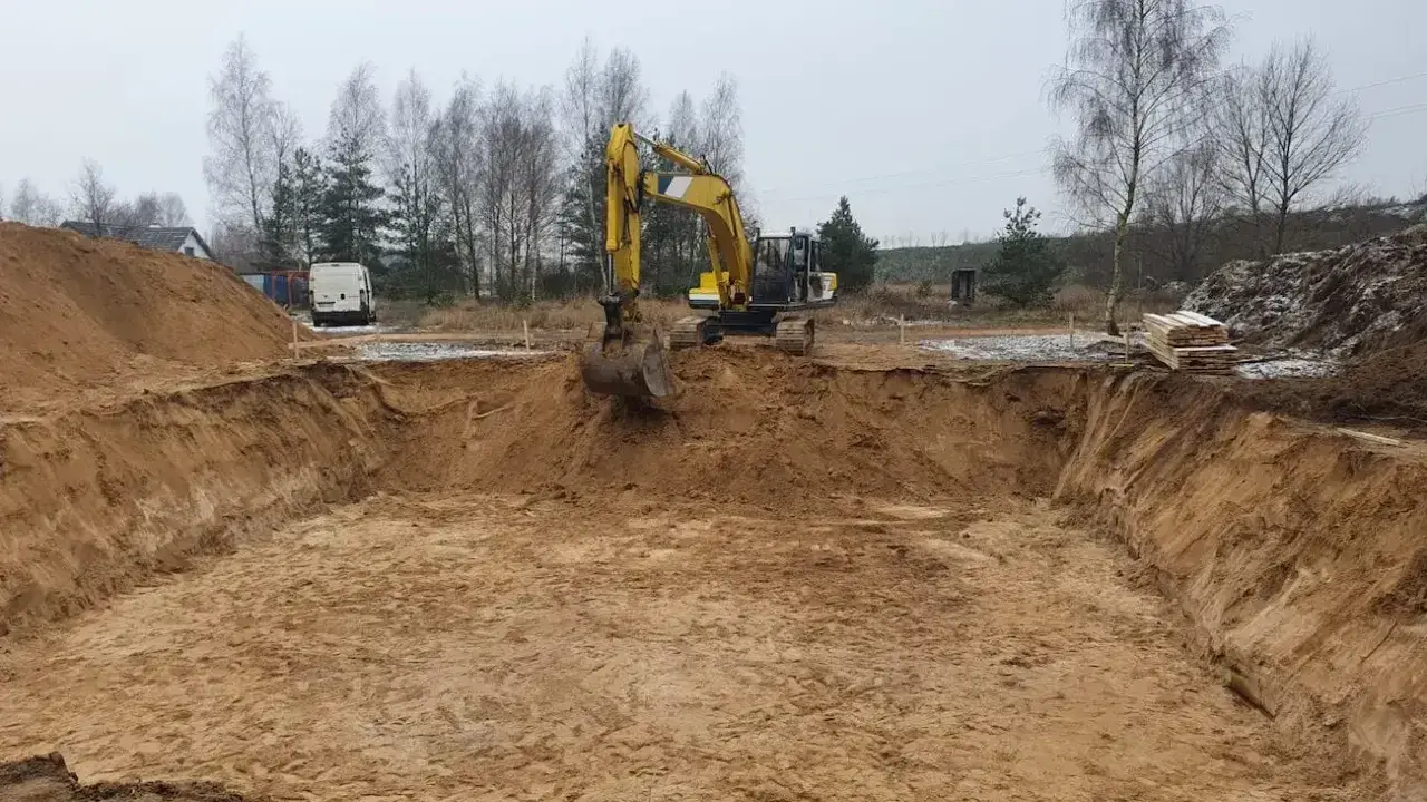 Koparka pracuje przy wykopy pod fundamenty. Widać głęboki dół w ziemi, usypane hałdy piasku i drzewa w tle.