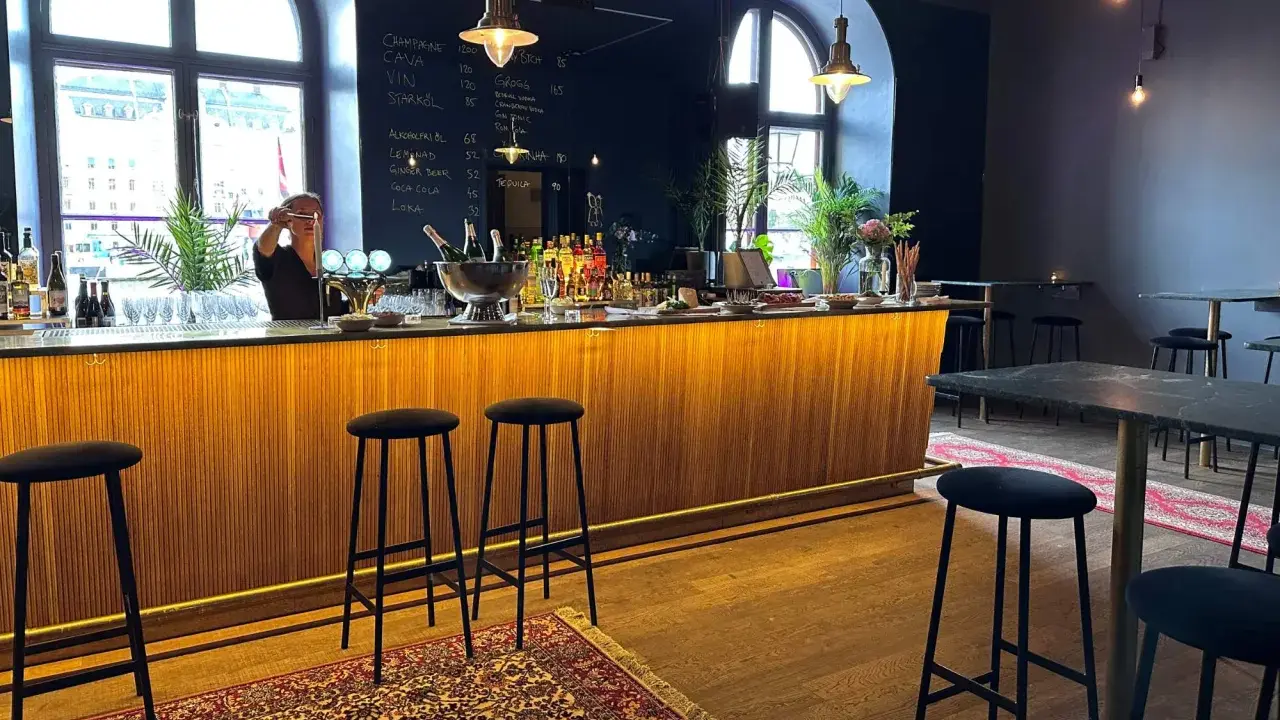 En bar med träpanel och svarta barstolar. En person bakom baren förbereder en drink. En meny på svarta tavlan visar priser för drycker i det musikaliska kvarteret.