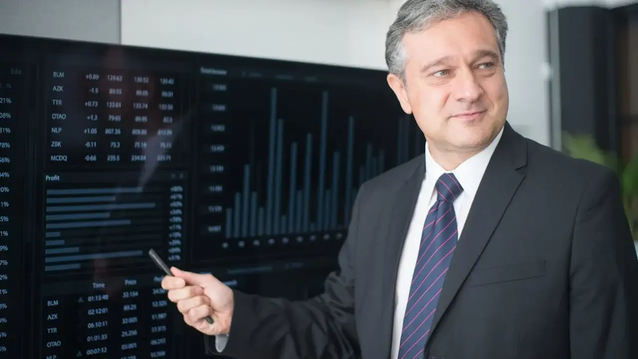 Mężczyzna w garniturze wskazuje na ekran z danymi finansowymi, wyjaśniając, kto płaci doradcy kredytowemu.