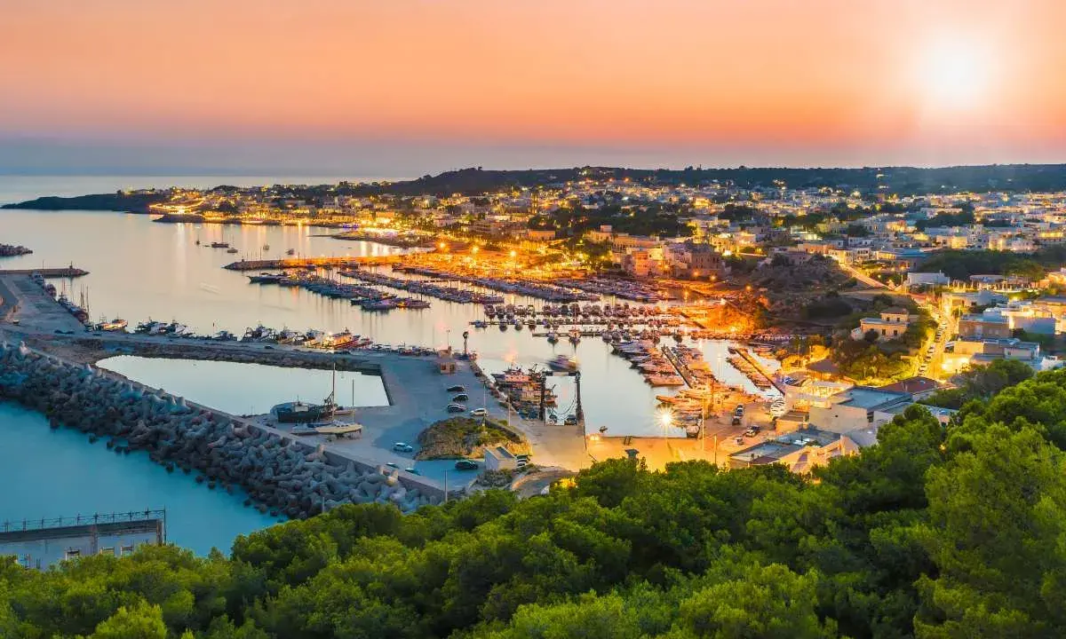 Porto illuminato al tramonto, un invito al turismo Salento. Barche ormeggiate e case che brillano sulla costa.