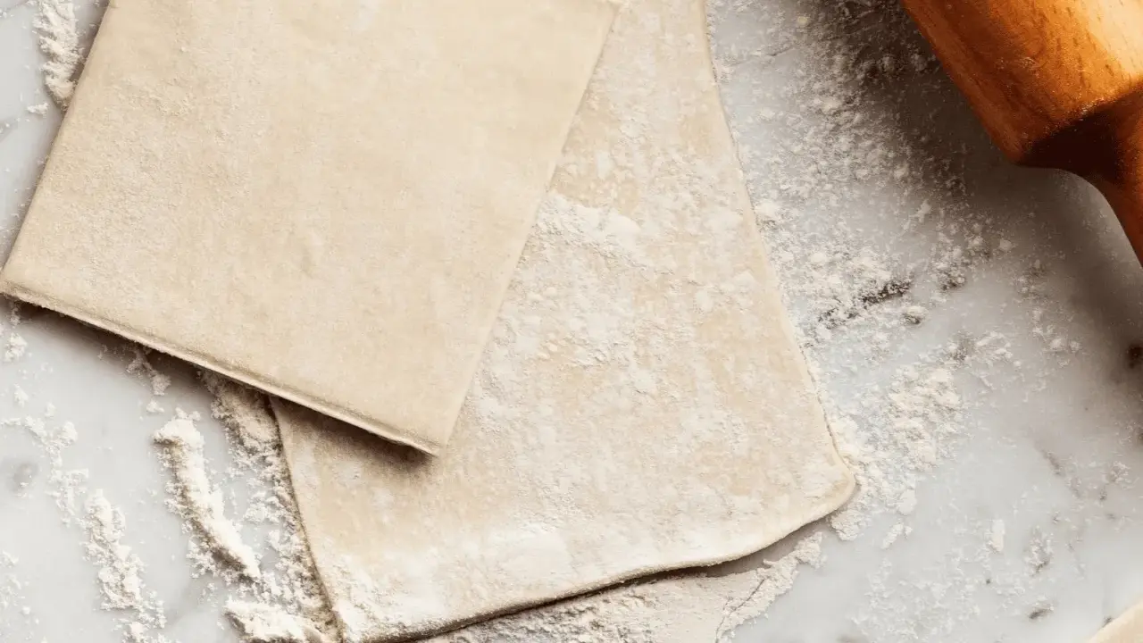 Pâte feuilletée prête à être travaillée, saupoudrée de farine sur un plan de travail en marbre. Un rouleau à pâtisserie en bois est visible sur la droite.