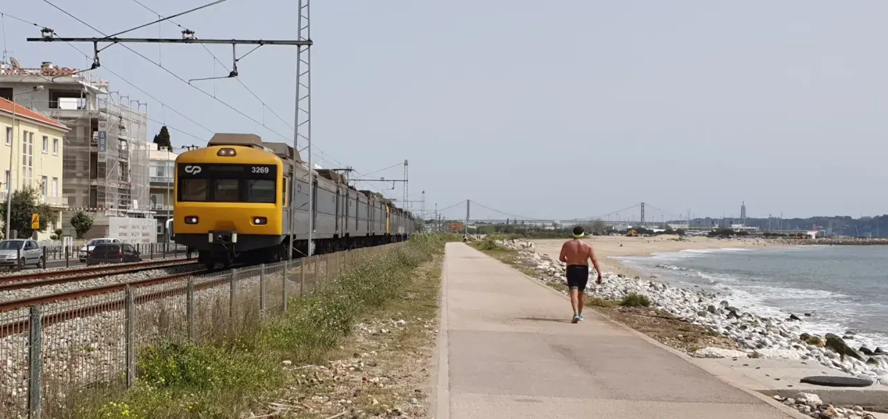 Gelber Zug fährt entlang der Küste von Cascais nach Lissabon. Ein Mann joggt am Strand entlang.