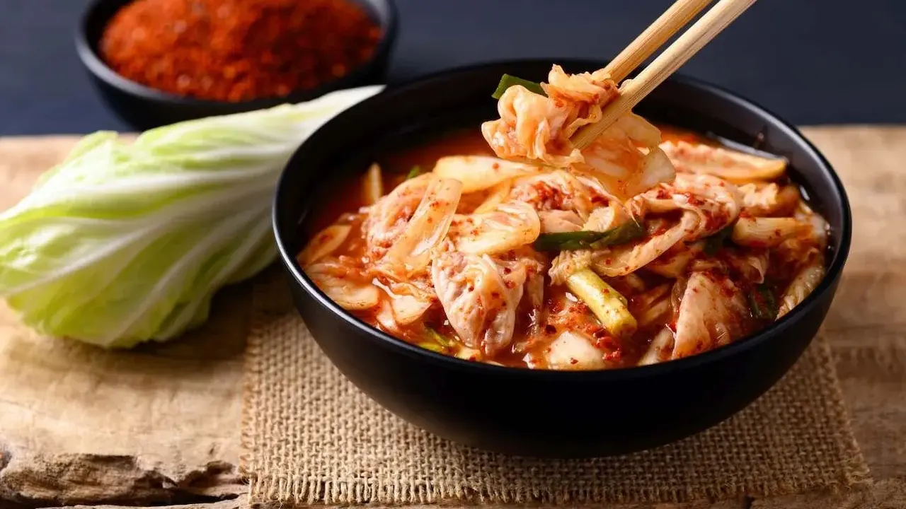 Miseczka z kimchi, pałeczki podnoszące kawałek kapusty, obok kapusta i miseczka z przyprawą.